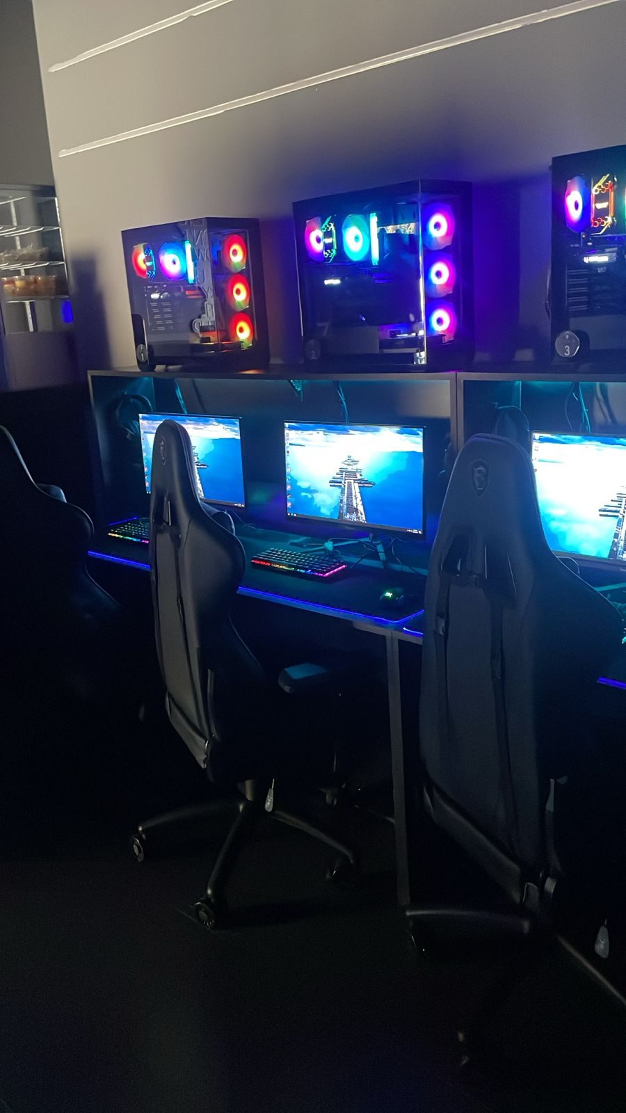 Three-station gaming setup with illuminated tower PCs on a shelf above monitors and racing chairs